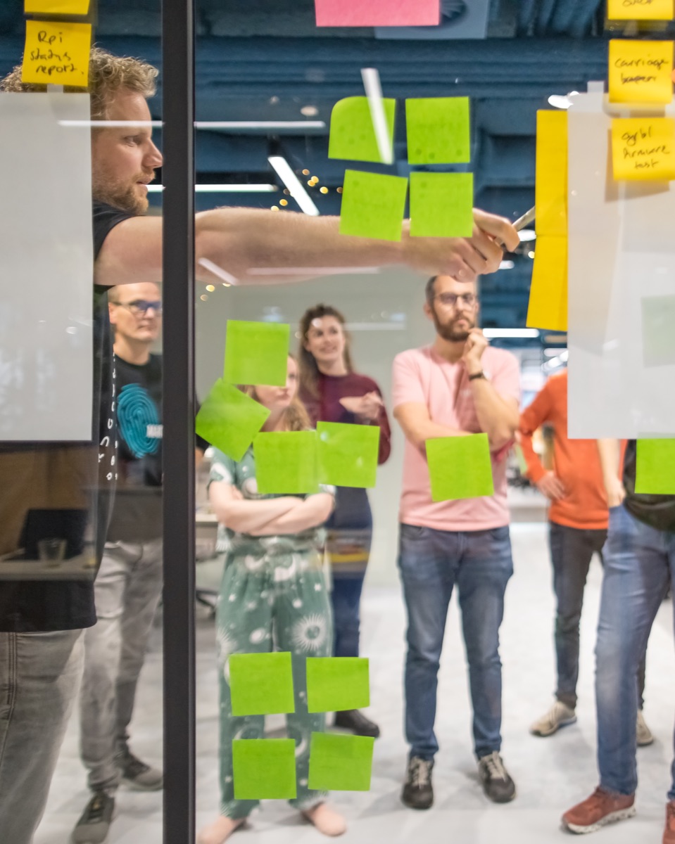 Deelnemers aan een hackathon verzamelen ideeën op post-its op een glazen wand